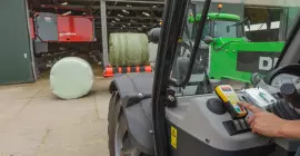 Activa una encintadora RW 1110 con sistema de control remoto frente a un establo con un robot alimentador en el fondo.