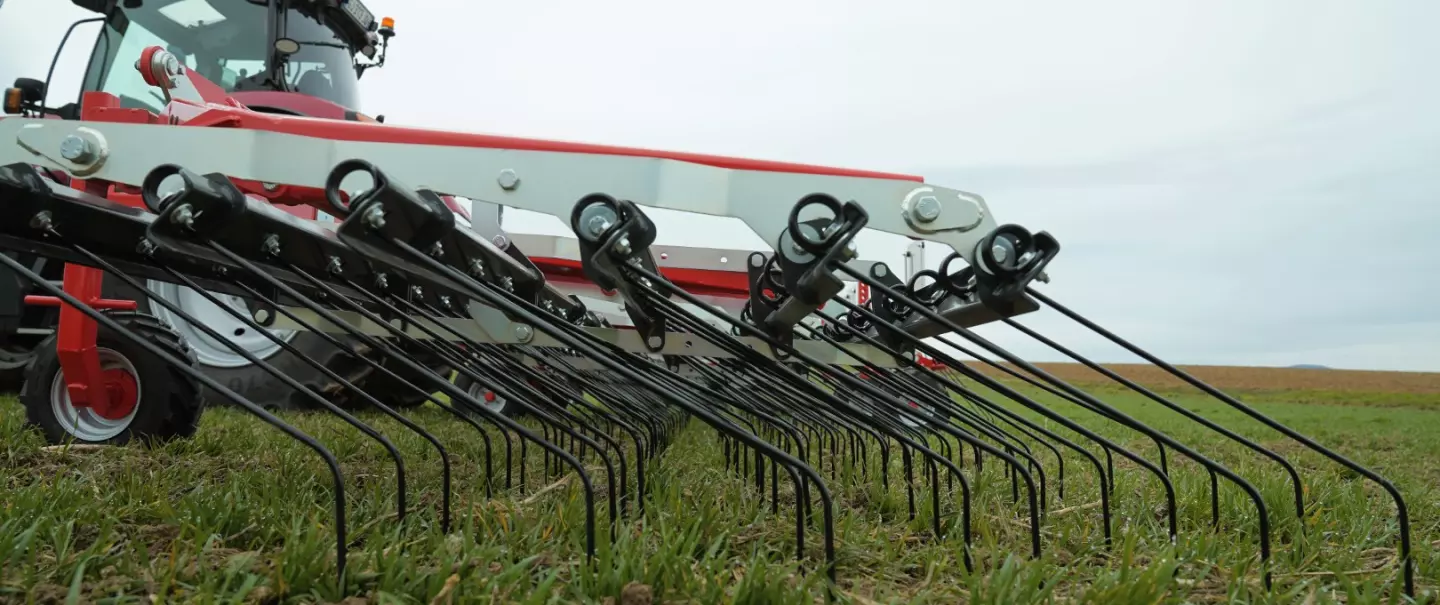 A red and white tractor with a tine weeder Tineliner is working in a grassy field under an overcast sky, preparing soil for farming.