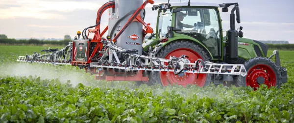 A green tractor with DELTIS mounted sprayer operates a large agricultural spraying implement across a field of green crops, creating visible spray mist under a cloudy sky.