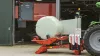 La RW 1110 encintando una paca redonda frente a un establo con un robot alimentador en el fondo.